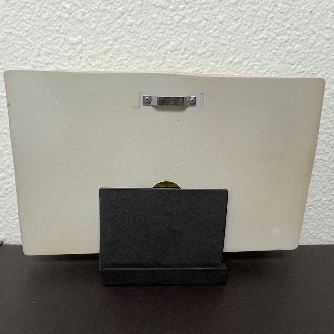 Back view of a wall plaque with black stand and mounting hardware against a textured wall