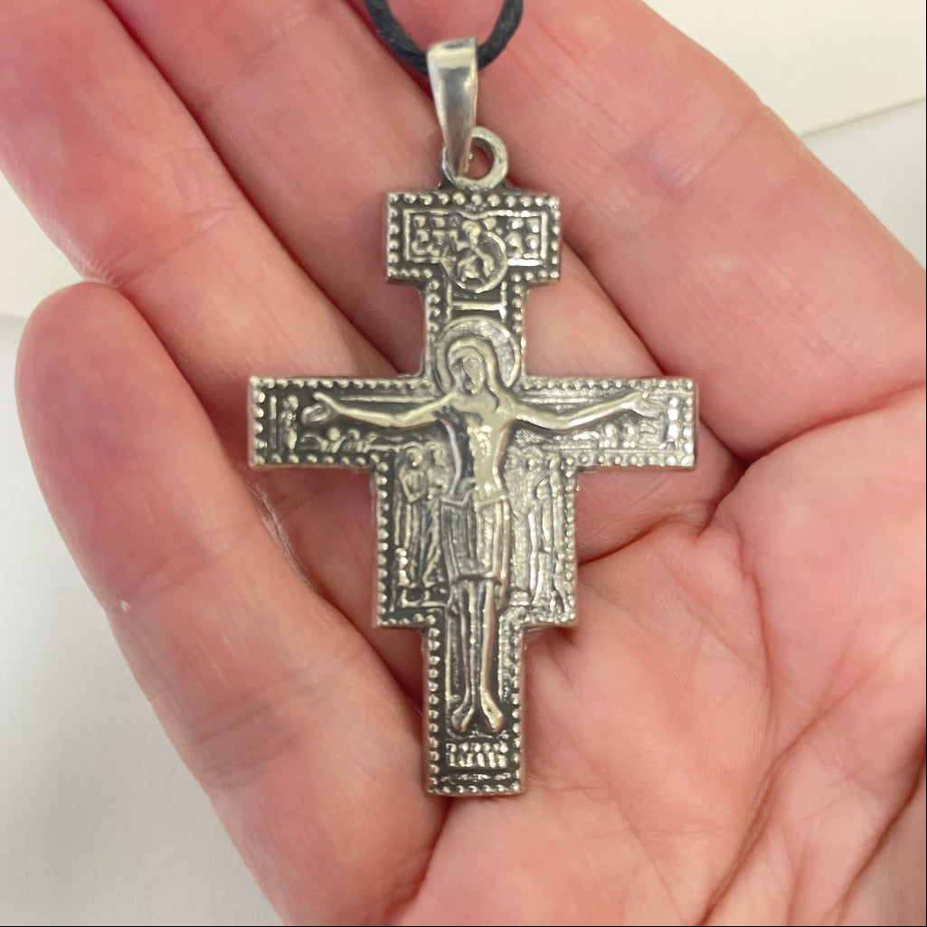 A silver-toned Orthodox cross pendant with an embossed figure of Jesus Christ, hanging from a black cord and held in a person's hand.