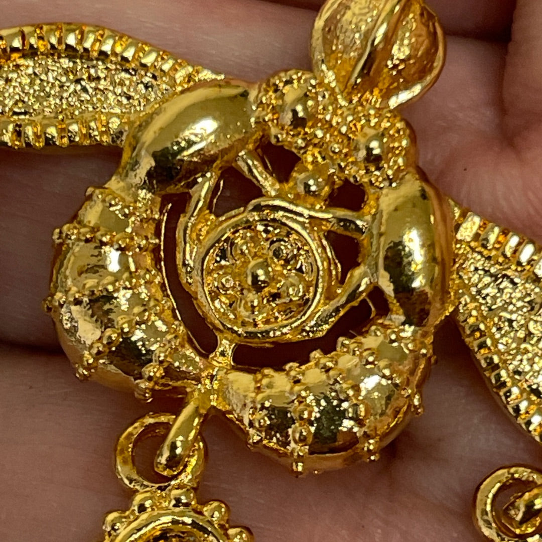 A close-up of a gold-toned ornate pendant or charm being held in a person's hand. The piece has intricate filigree and textured beaded details, with a circular medallion-like center and a hanging loop at the bottom.