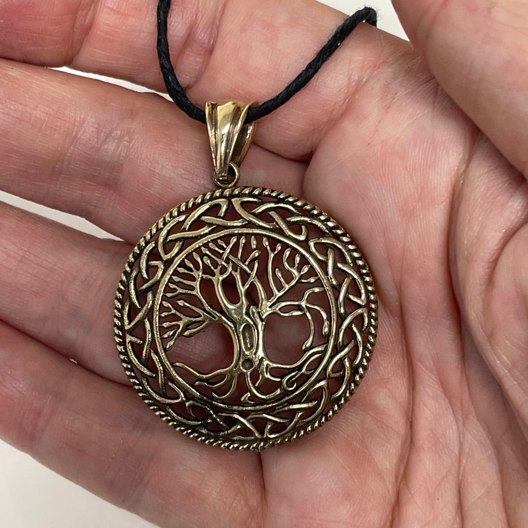 A circular tree of life pendant with Celtic knotwork patterns, held in a hand. The pendant has a textured antique bronze finish and hangs from a black cord.
