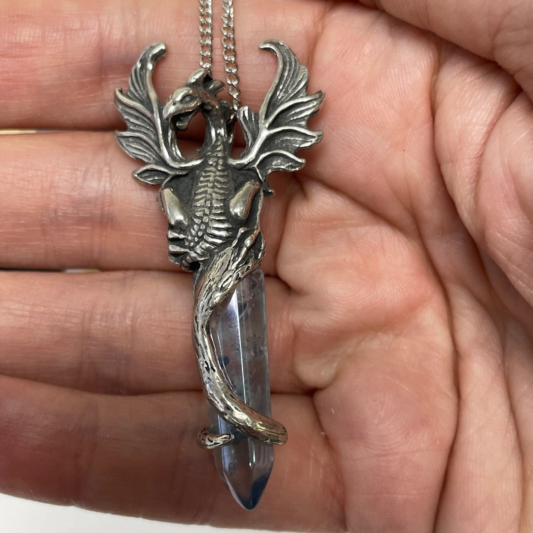 A hand holding a detailed silver dragon pendant with a clear blue-gray crystal point at the bottom.