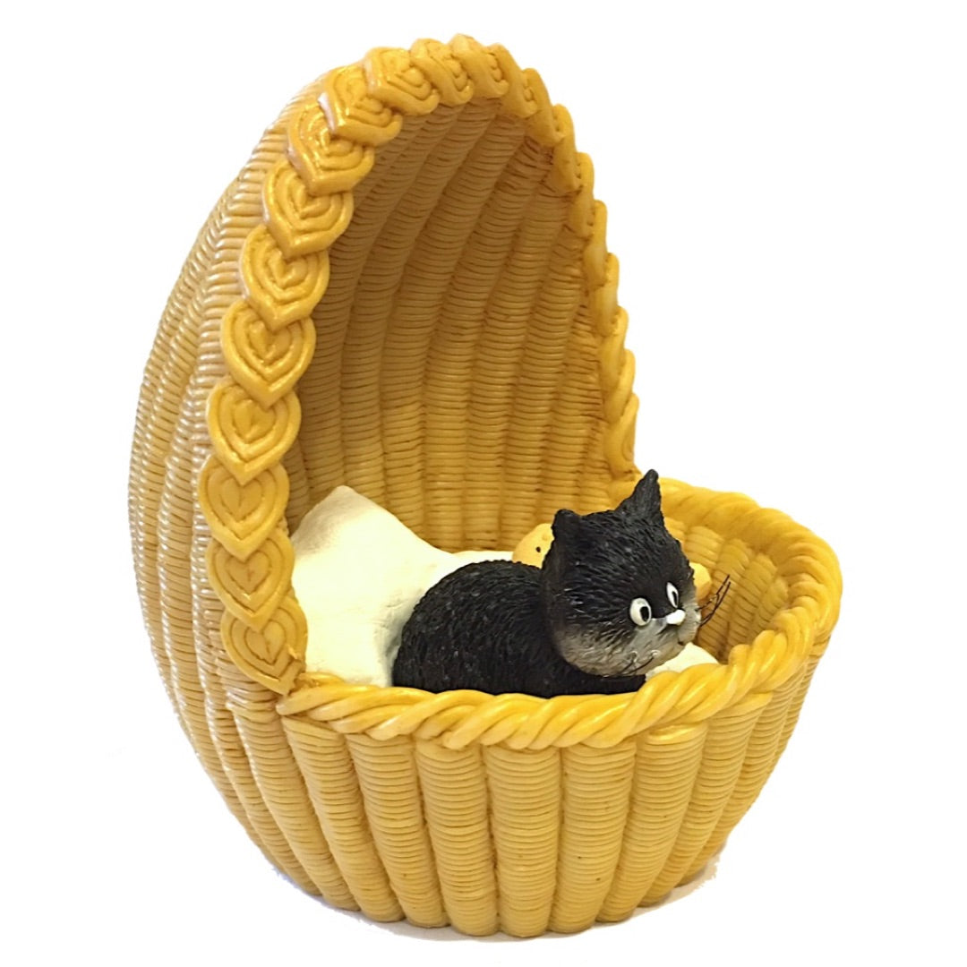 A small black cat figurine resting inside a yellow woven basket cradle with a white cushion, photographed on a plain white background.