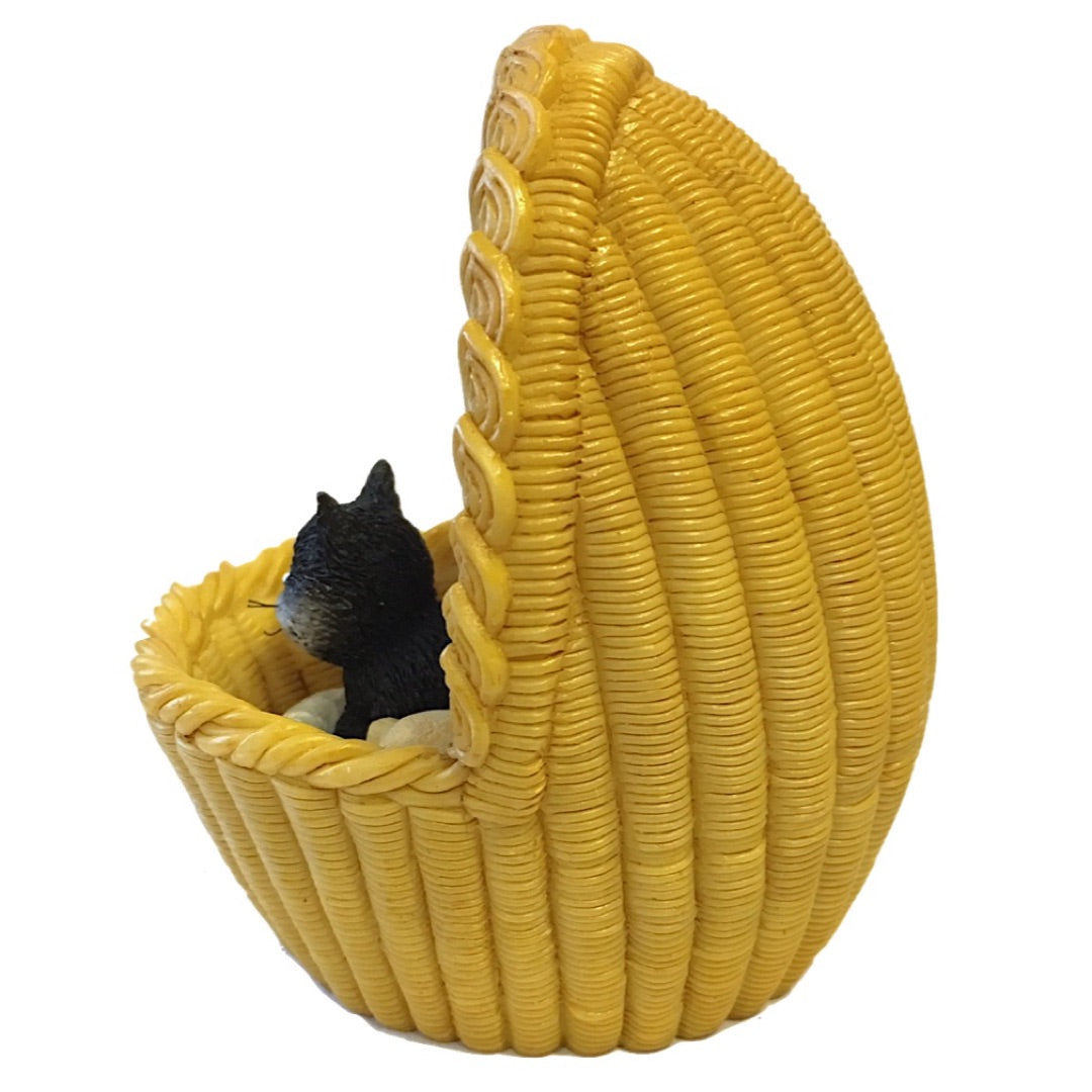 A small black cat figurine sitting inside a yellow woven basket-style pet tower or perch, photographed on a plain white background.