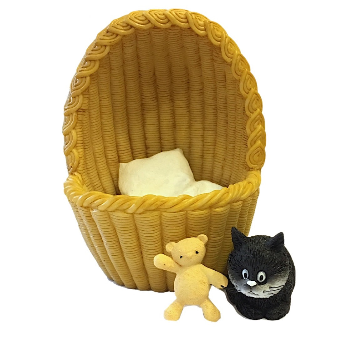 A yellow woven basket chair with a white cushion, accompanied by a small yellow teddy bear and a black cat figurine on a plain white background.