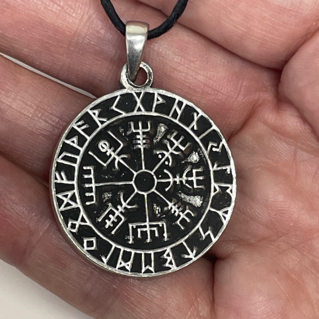 A silver-toned circular pendant with engraved runic symbols and a central compass-like motif, hanging from a black cord and held in a person's hand.