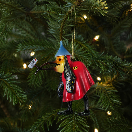 A Christmas ornament shaped like a bird wearing a red cape and blue hat, hanging on a decorated Christmas tree with warm lights.