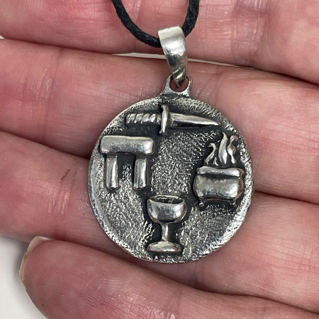 A silver-toned circular pendant necklace with a black cord, held in a person's hand. The pendant features embossed religious or ceremonial symbols including a dagger, a chalice, and a cauldron.