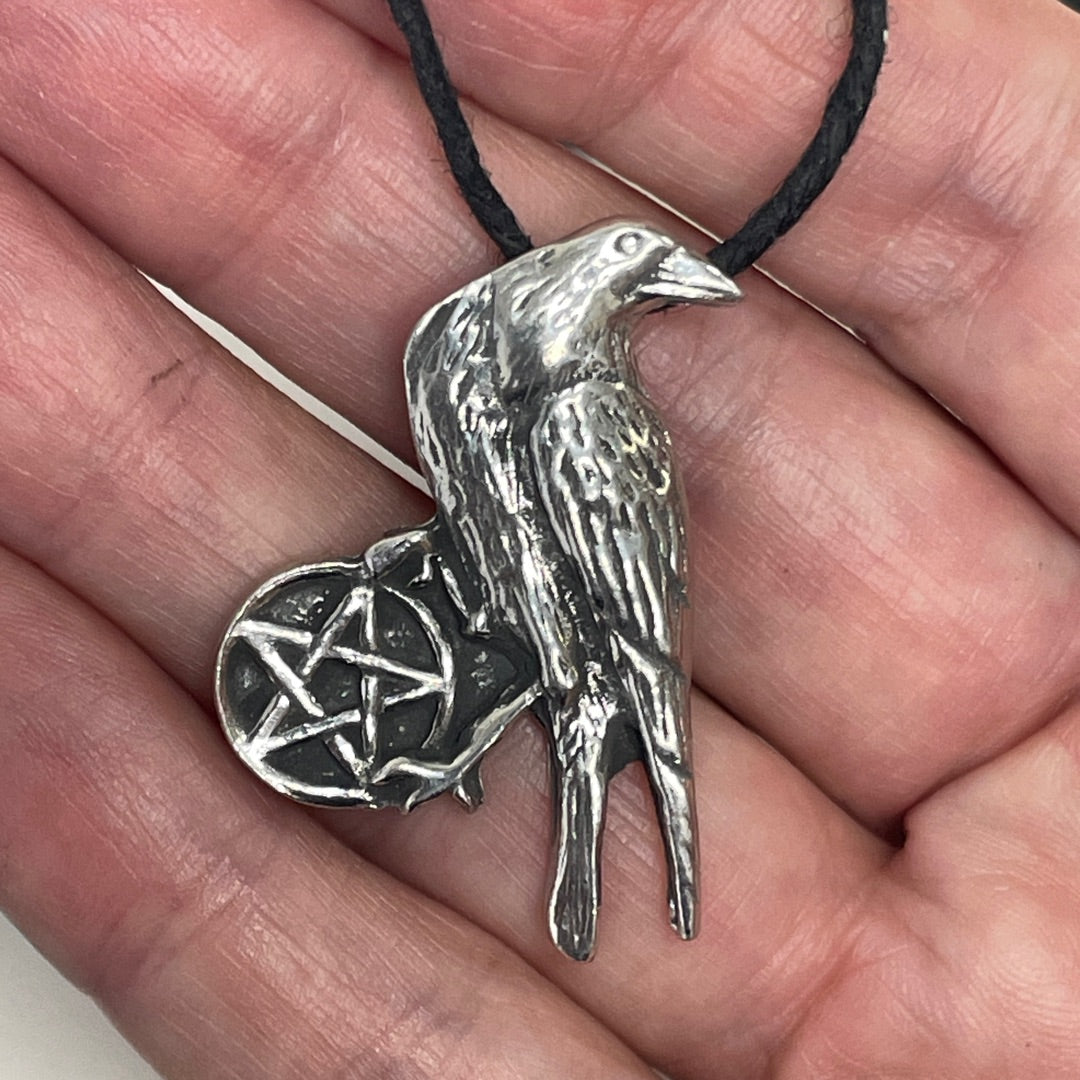 A silver raven pendant necklace with a pentagram design, held in a person's hand on a black cord.